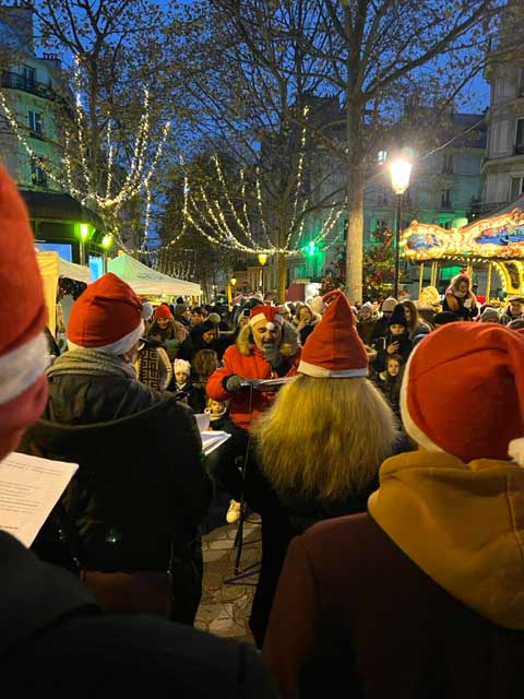 Les membres de la chorale Envie de chanter pour la fête de Noël, place Lino Ventura dans le 9ème à Paris. On voit les illuminations aux arbres.
