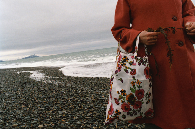 Photo de Françoise Huguier représentant une femme au manteau rouge dont on ne voit pas le visage, portant un sac, se promenant sur un plage de galets.