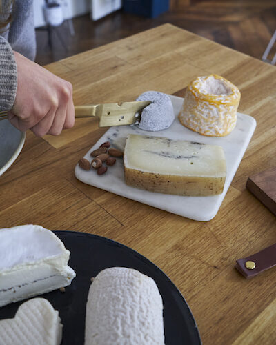 Gros plan sur une main qui coupe un fromage de chèvre sur un plateau, avec d'autres fromages autour.
