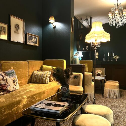Salon de l'hôtel La Mondaine : canapé et poufs de velours jaune, mur sombre; gravures au mur et ambiance tamisée.