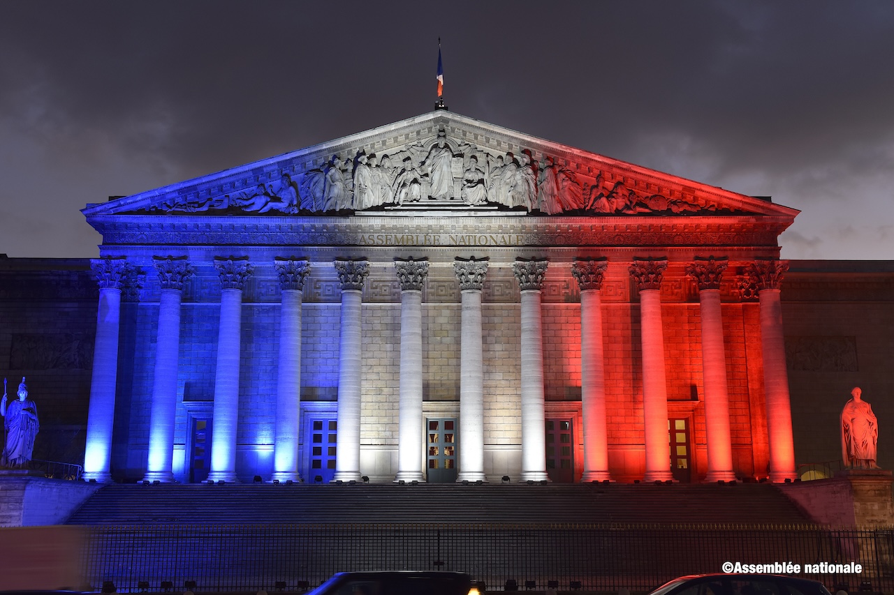 Photo des colonnades de l'Assemblée nationale éclairée de bleu blanc rouge