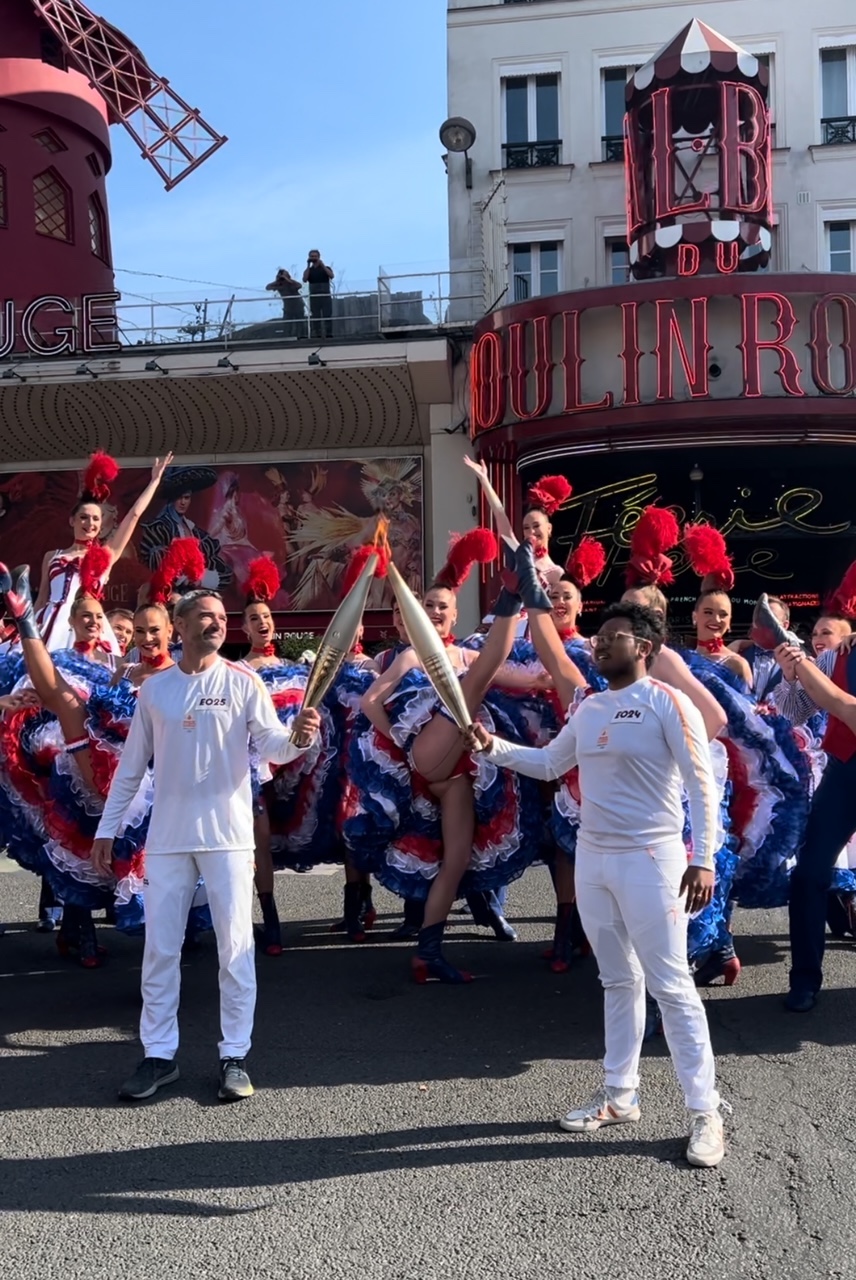 Les deux porteurs s'échangent la flamme devant le Moulin Rouge alors que les danseuses dansent le French cancan